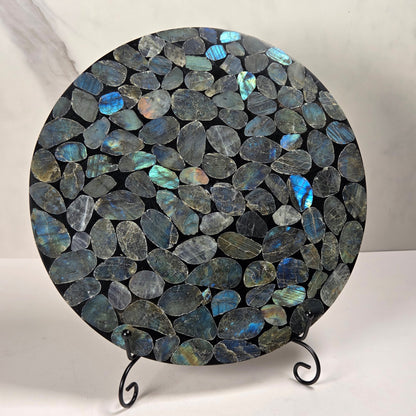 A round decorative board or tray covered in polished irregular labradorite stones with blue, green, and gray flashes, displayed on a small black metal stand against a light marble-patterned background.
