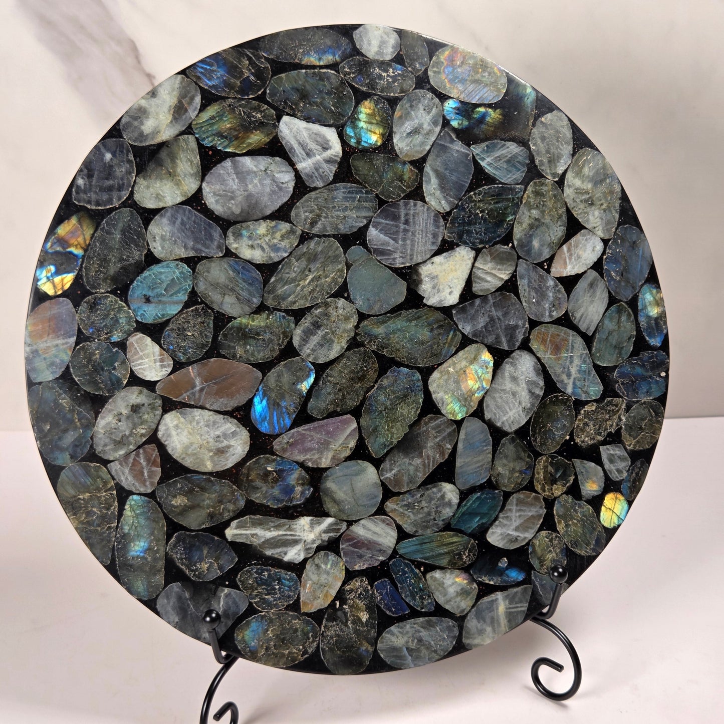 A round decorative board or tray covered in polished irregular labradorite stones with blue, green, and gray flashes, displayed on a small black metal stand against a light marble-patterned background.