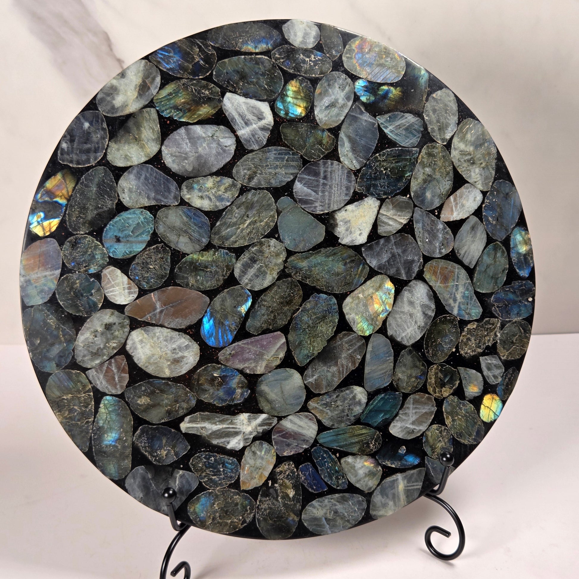 A round decorative board or tray covered in polished irregular labradorite stones with blue, green, and gray flashes, displayed on a small black metal stand against a light marble-patterned background.