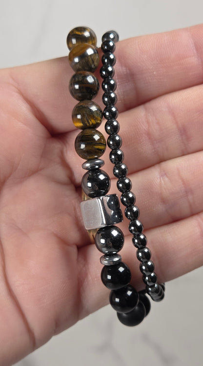 Two beaded stretch bracelets held in a hand against a light gray marble background. One bracelet features faceted brown and black tiger's eye beads with a silver spacer, and the other features small dark gray and black faceted beads.