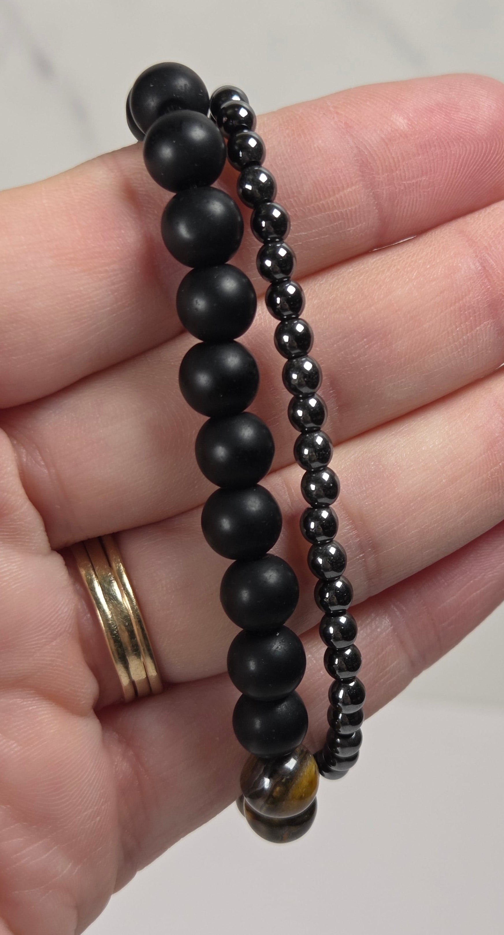 Two beaded stretch bracelets in black, one with larger matte round beads and the other with smaller glossy round beads, held in a person's hand.