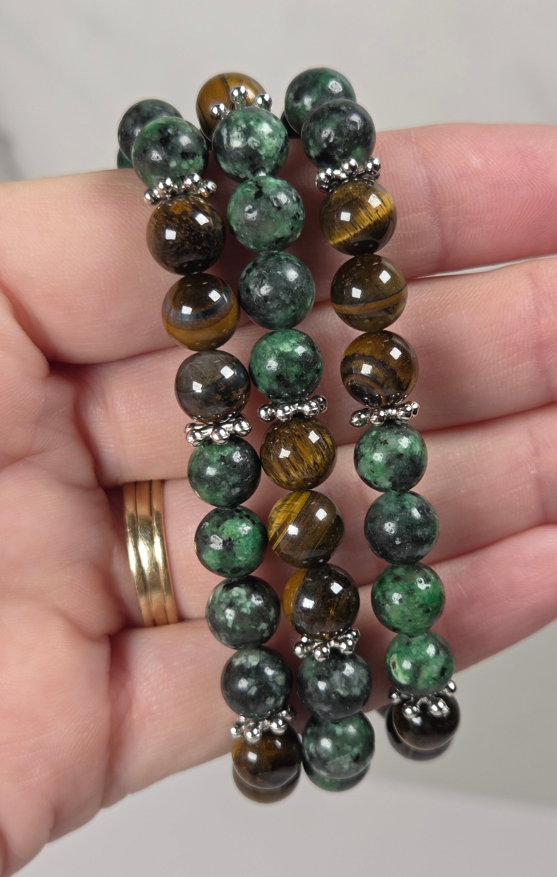 Three beaded stretch bracelets held in a hand. The bracelets feature round beads in green malachite, brown tiger's eye, and dark gray obsidian, separated by small silver spacer beads.