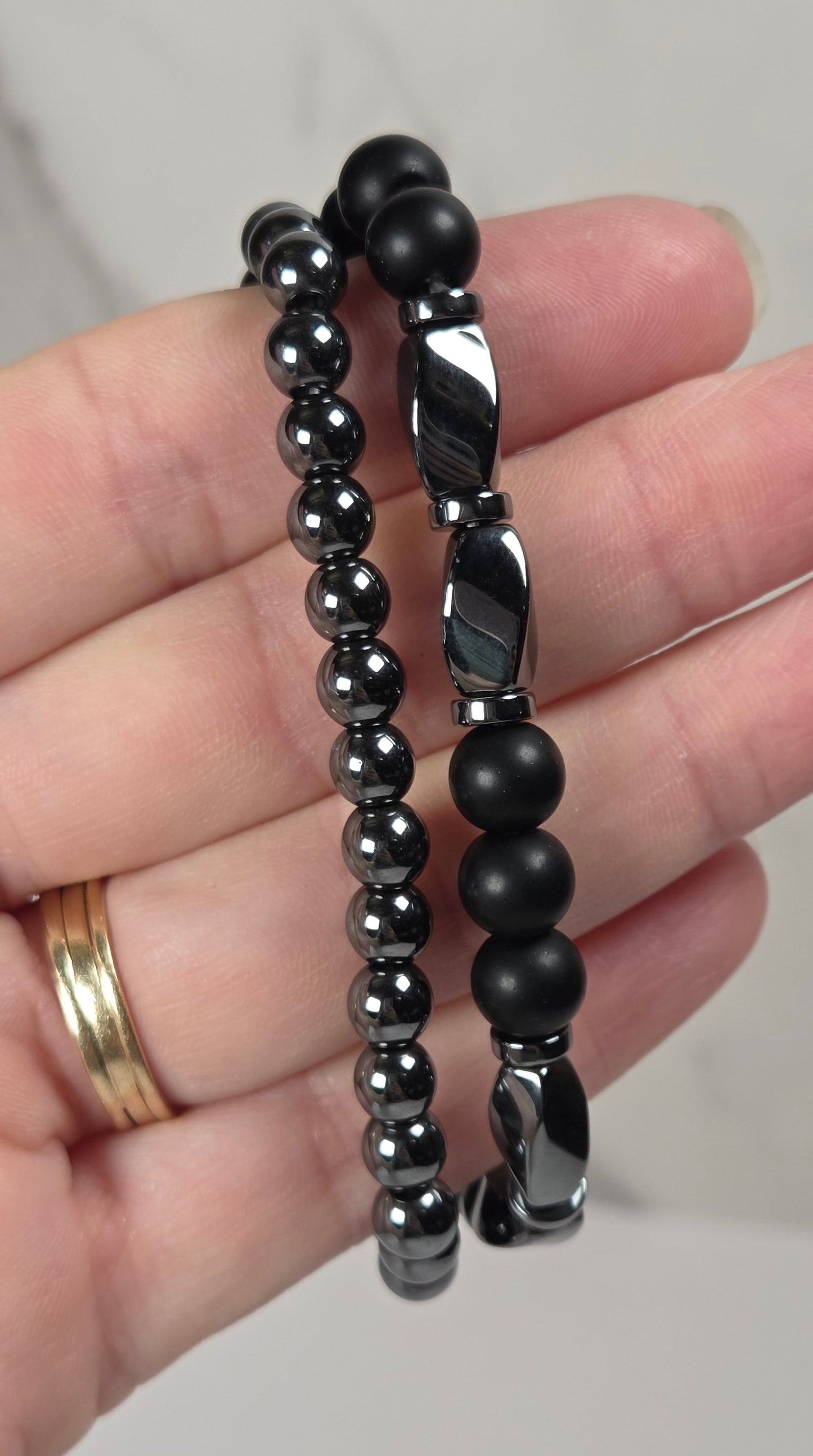 Two black beaded bracelets, one with smooth matte beads and the other with polished metallic beads, held in a hand against a light gray marble background.