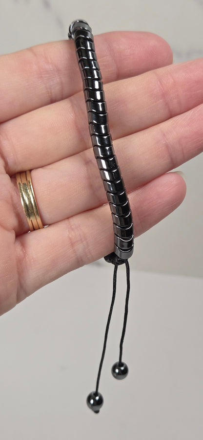 A black segmented adjustable bracelet held in a person's hand, featuring two dangling black beads on a light gray background.