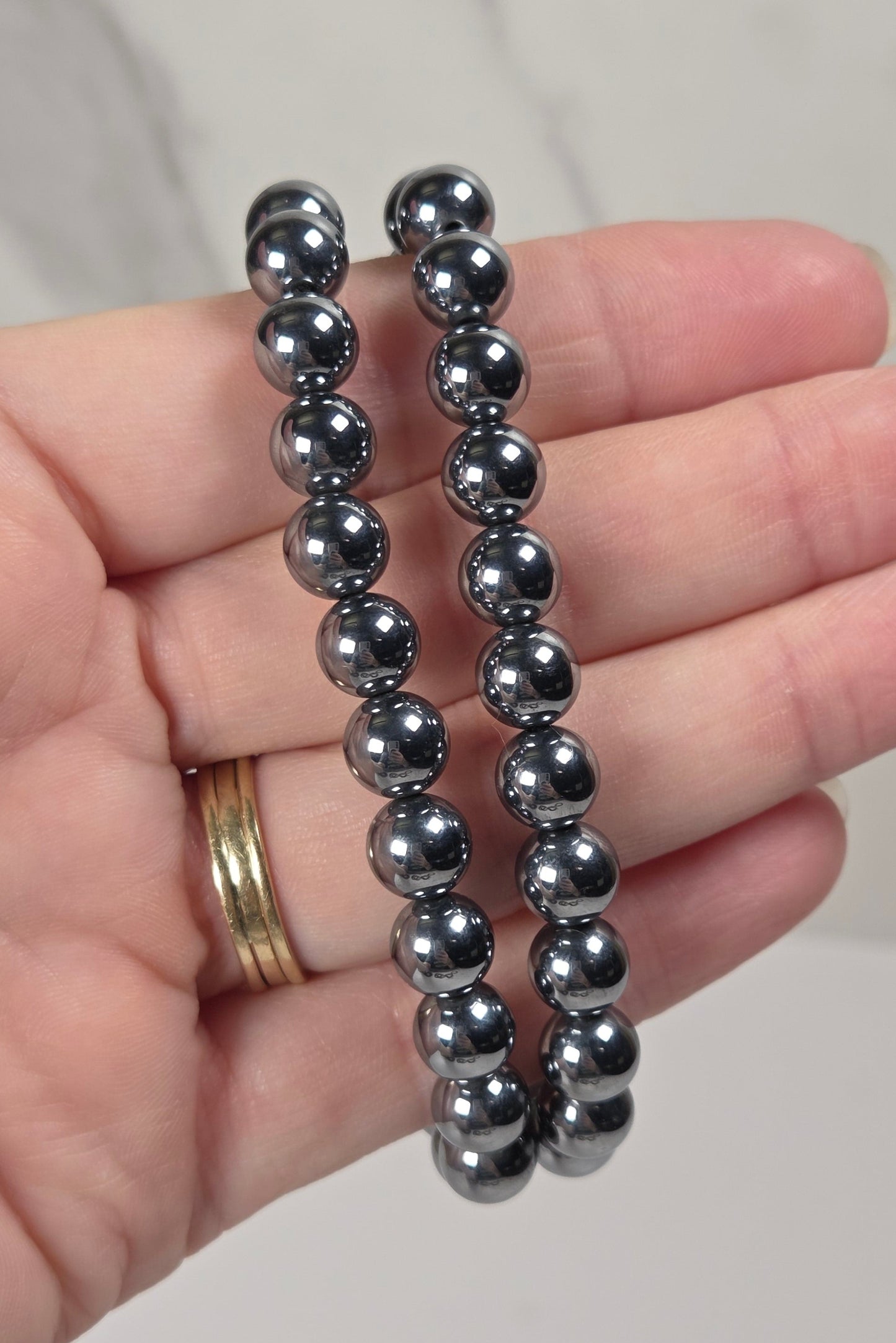 Two dark metallic beaded bracelets held in a person's hand against a light marble background.
