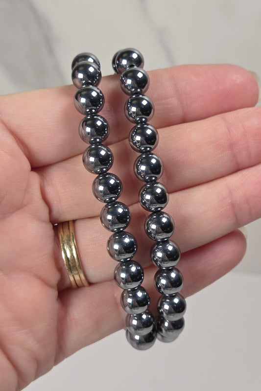 Two dark metallic beaded bracelets held in a person's hand against a light marble background.