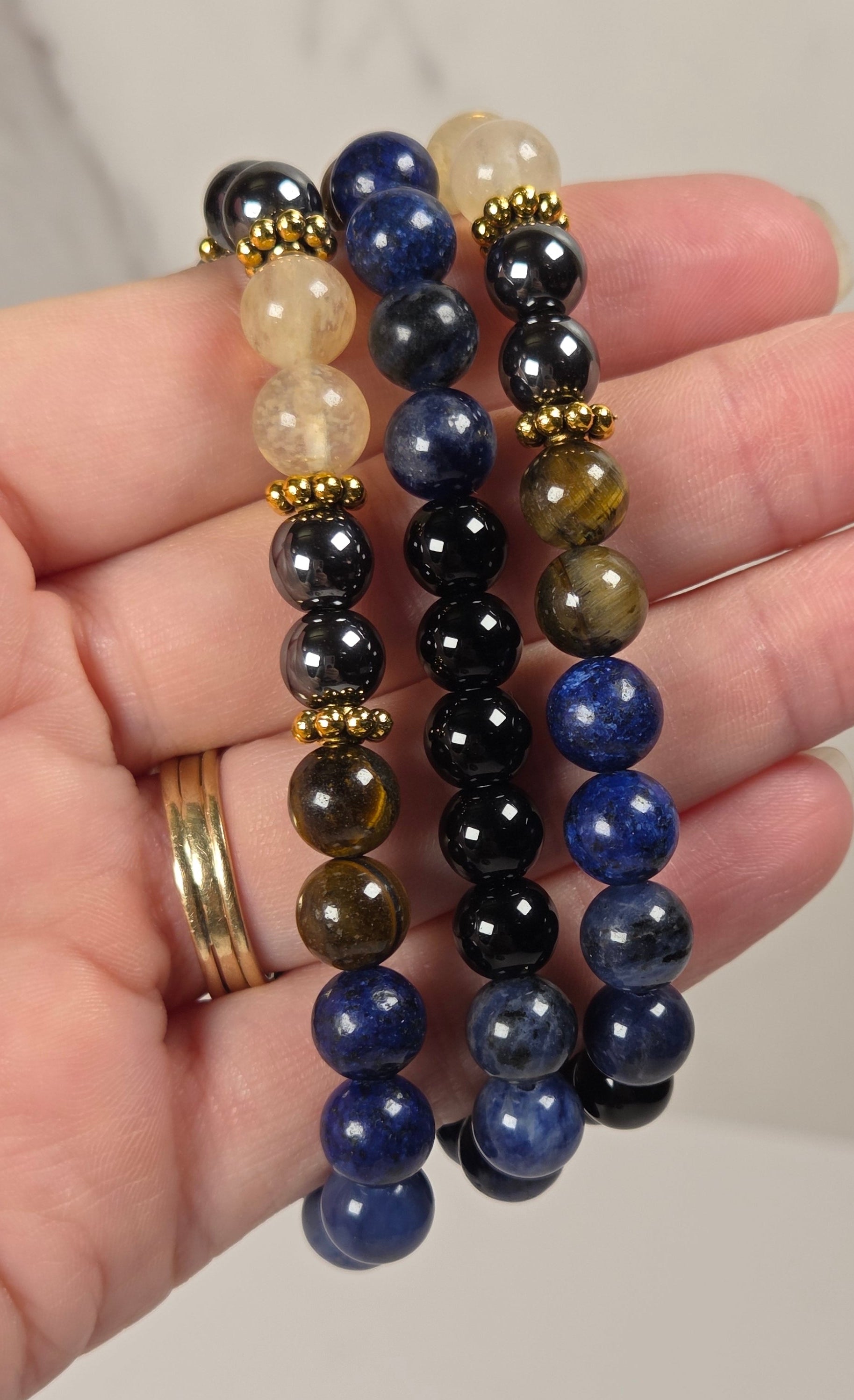 Three beaded stretch bracelets held in a hand. The bracelets feature polished round beads in navy blue, black, translucent cream, and tiger-eye patterns, separated by small gold spacer beads.