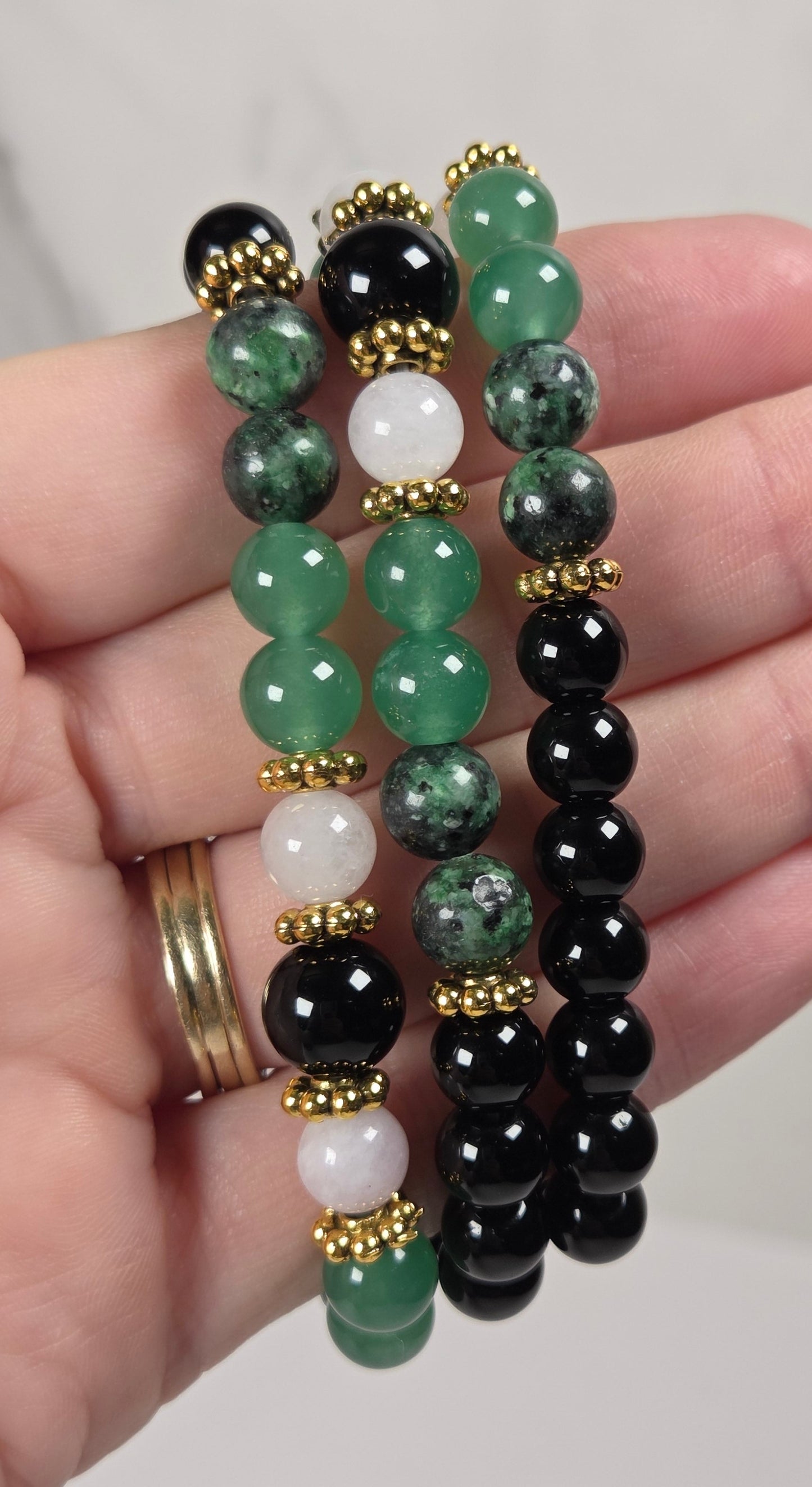 Three beaded stretch bracelets held in a hand. The bracelets feature polished round beads in black, various shades of green, and white, separated by small gold spacers.