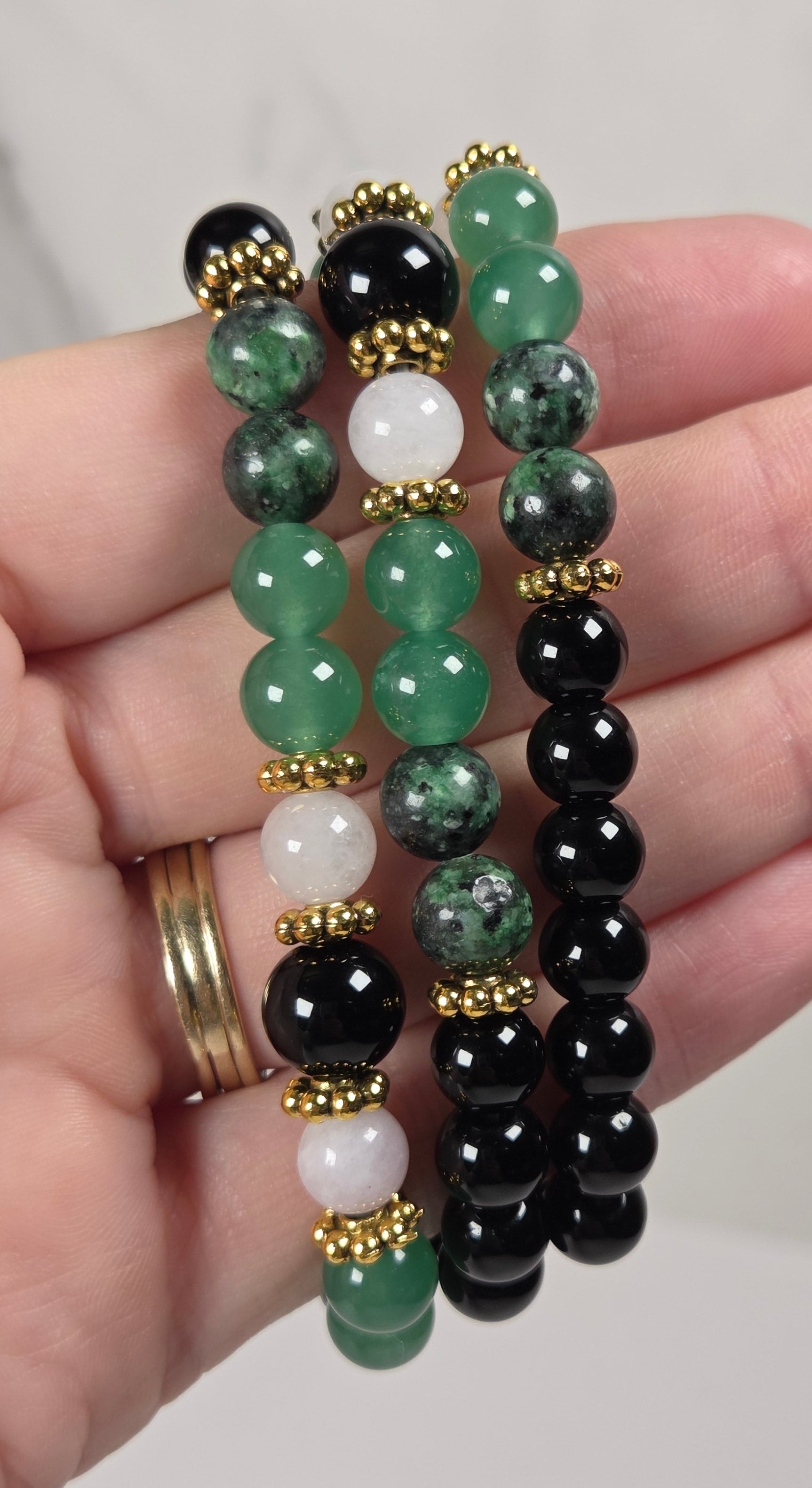 Three beaded stretch bracelets held in a hand. The bracelets feature polished round beads in black, various shades of green, and white, separated by small gold spacers.