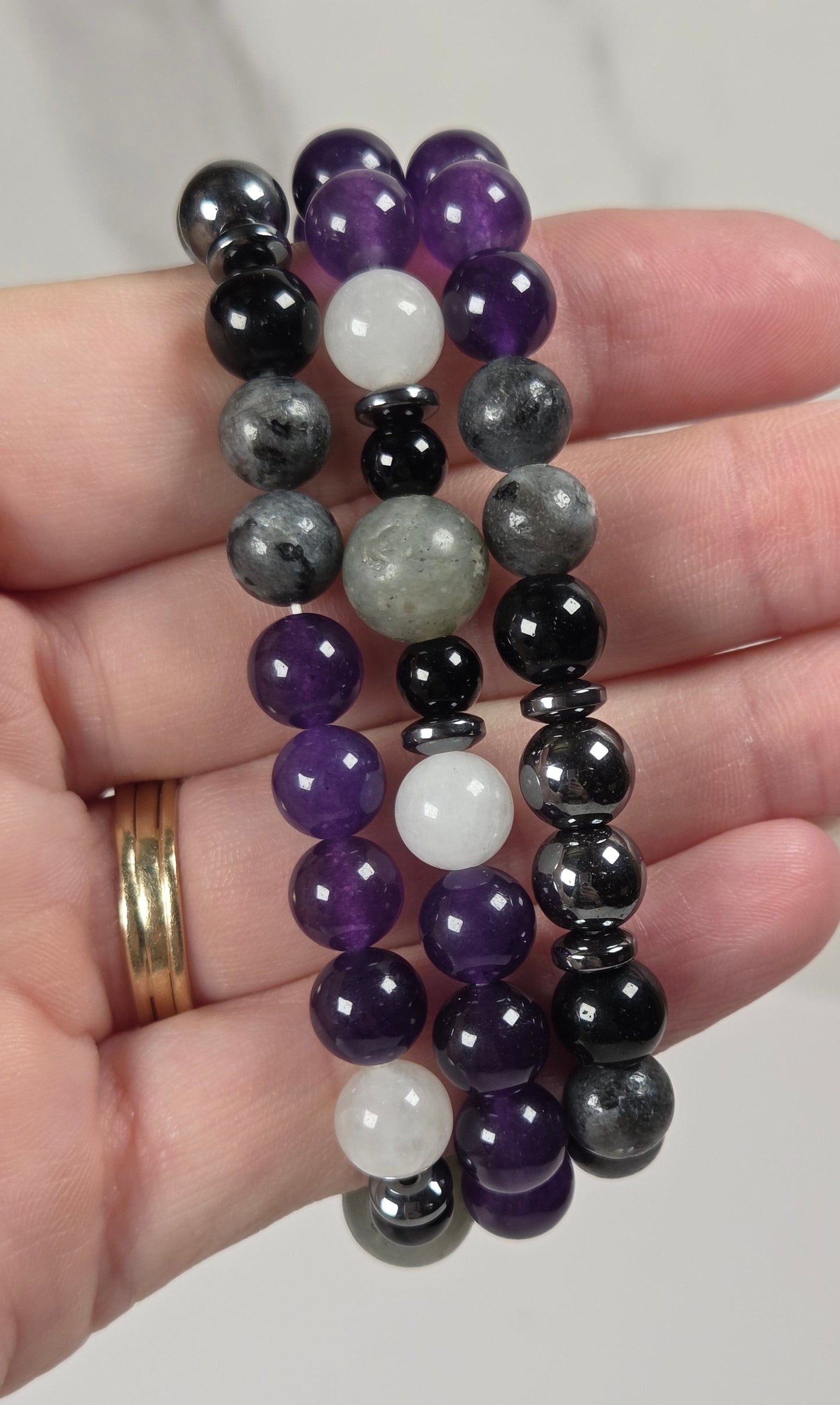 Three beaded stretch bracelets in purple, black, gray, and white stone, held in a person's hand against a light gray marble background.