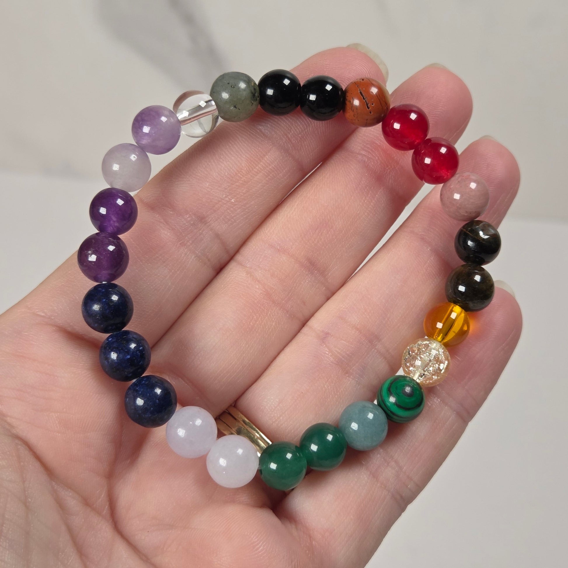 A multicolor beaded stretch bracelet made of polished gemstone-like beads, held in a person's hand. The bracelet features a rainbow sequence of beads including purple, blue, green, yellow, orange, red, black, and white, with a small clear faceted bead near the bottom.