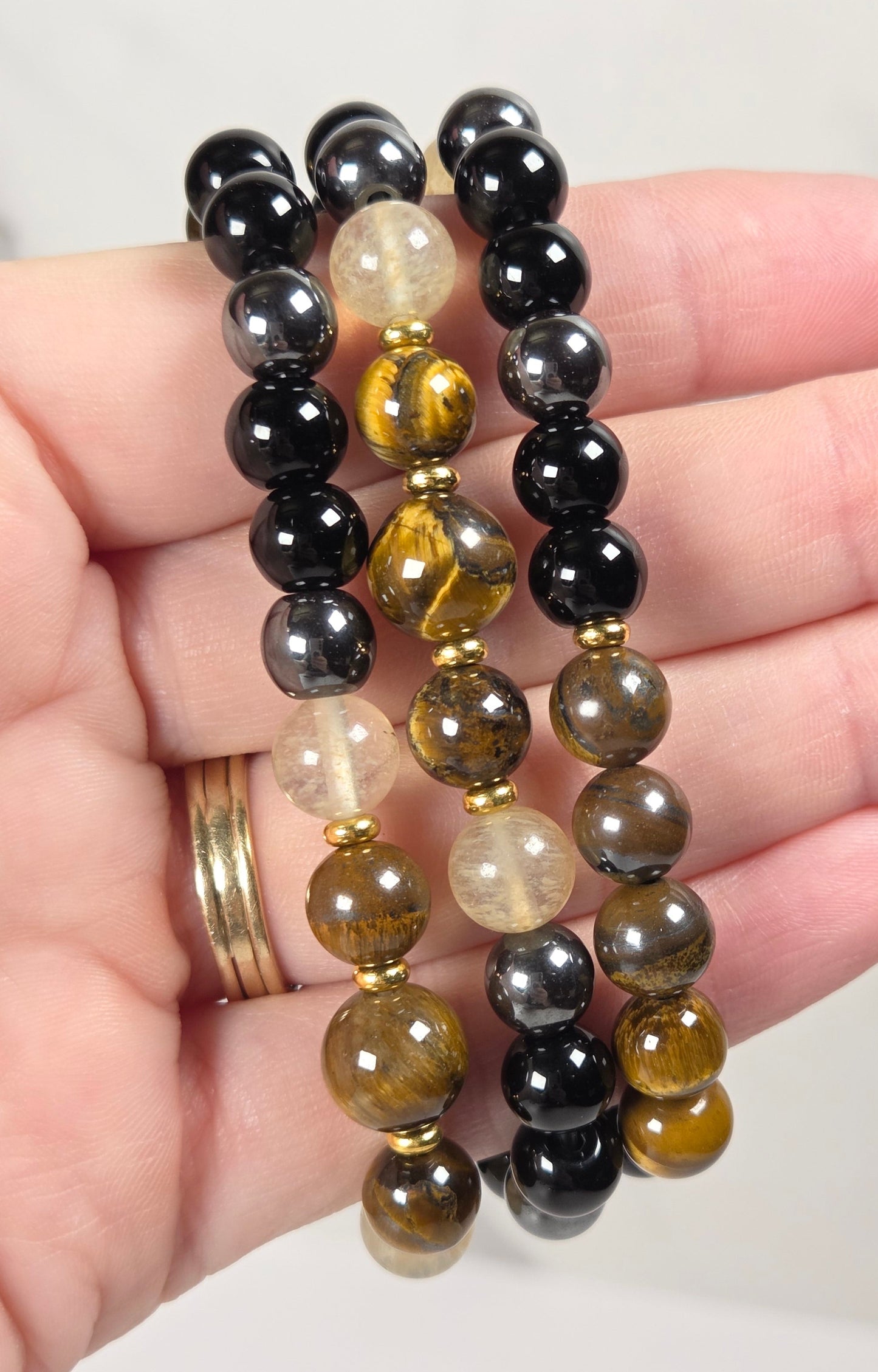 Three beaded stretch bracelets held in a hand. The bracelets feature polished round beads in black, metallic gold, brown, and translucent milky white, separated by small gold spacer beads.
