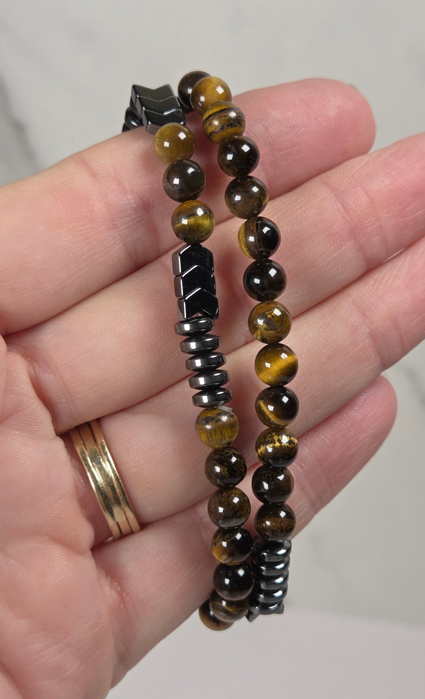 A close-up of a hand holding two beaded bracelets. One bracelet features polished round beads in brown, black, and golden-yellow tiger's eye patterns, while the other is made of dark metallic spacer beads.