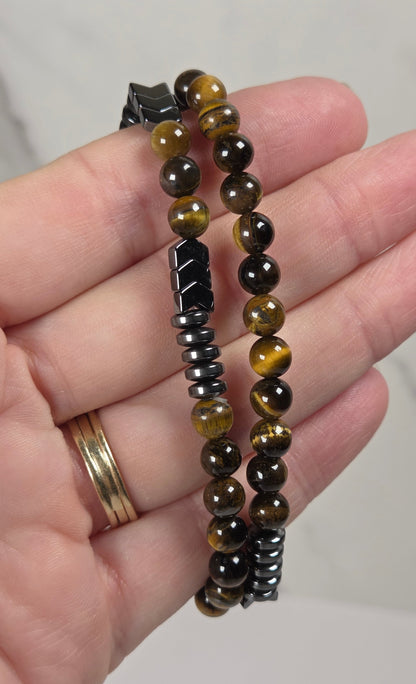 A close-up of a hand holding two beaded bracelets. One bracelet features polished round beads in brown, black, and golden-yellow tiger's eye patterns, while the other is made of dark metallic spacer beads.