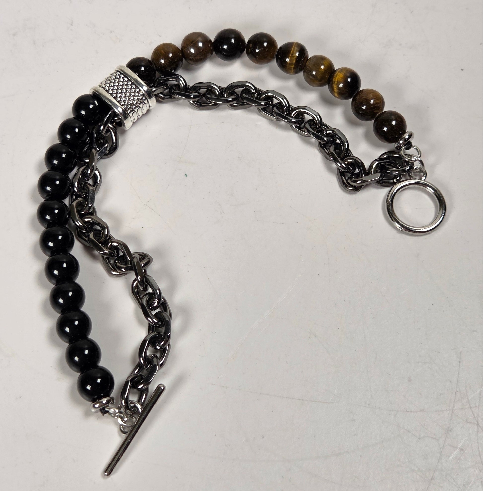 A mixed-material beaded and chain bracelet displayed on a light gray surface. The bracelet features black faceted beads, brown tiger's eye beads, a textured silver spacer, a dark gunmetal chain, and a silver toggle clasp.