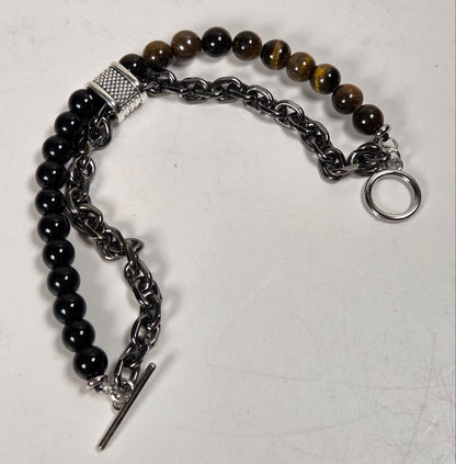 A mixed-material beaded and chain bracelet displayed on a light gray surface. The bracelet features black faceted beads, brown tiger's eye beads, a textured silver spacer, a dark gunmetal chain, and a silver toggle clasp.