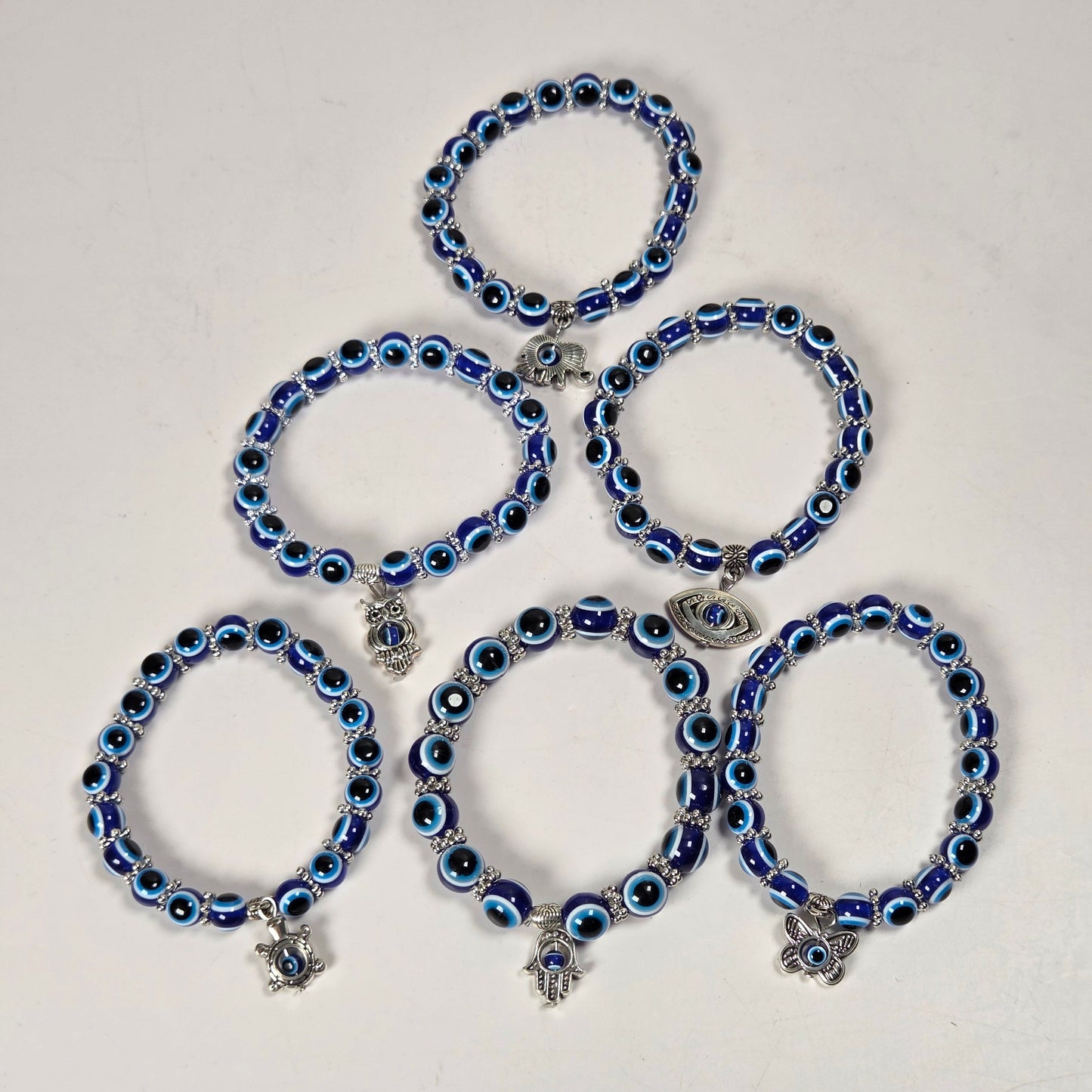 Five blue and black evil eye beaded bracelets with silver charms, arranged in a pyramid on a plain off-white background.