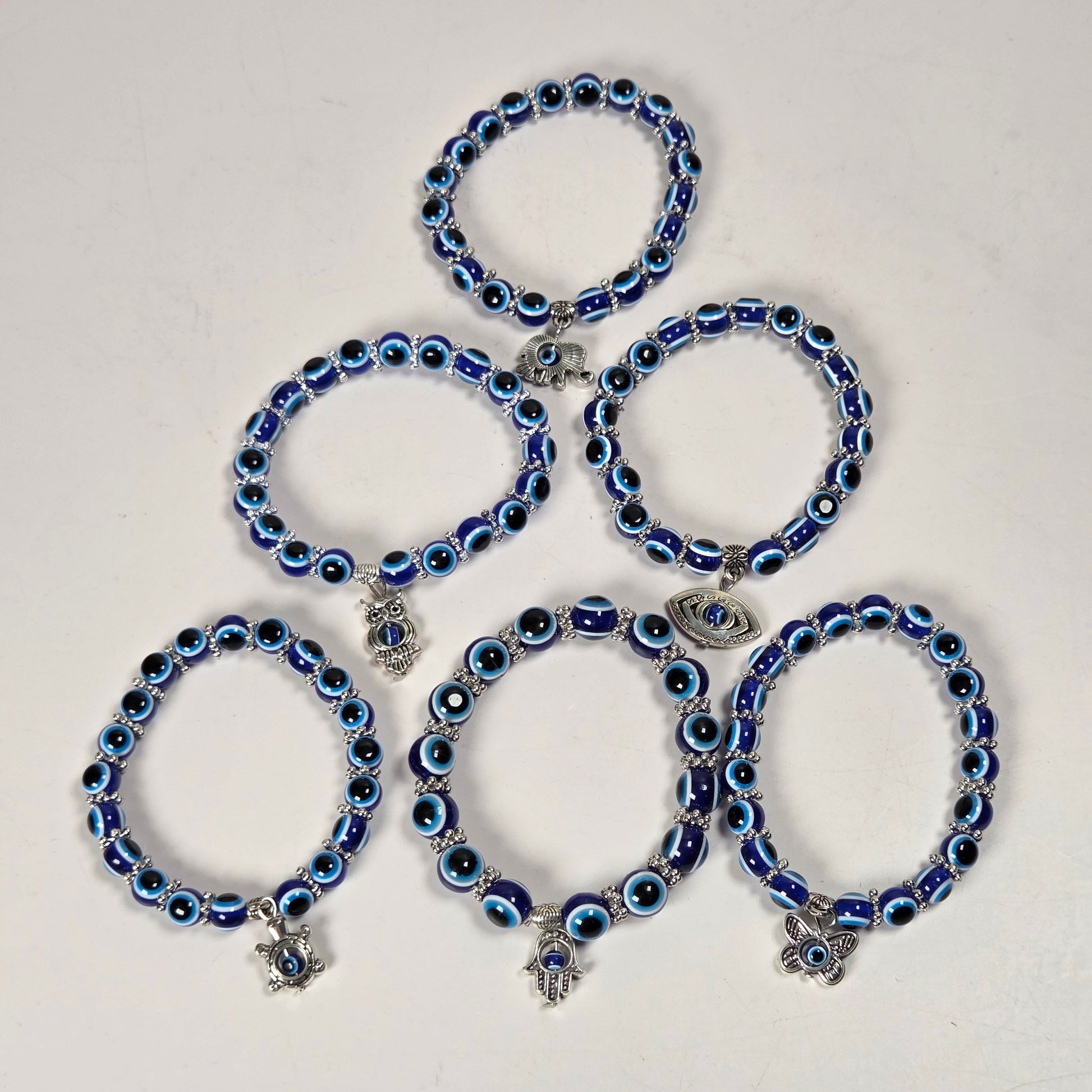 Five blue and black evil eye beaded bracelets with silver charms, arranged in a pyramid on a plain off-white background.