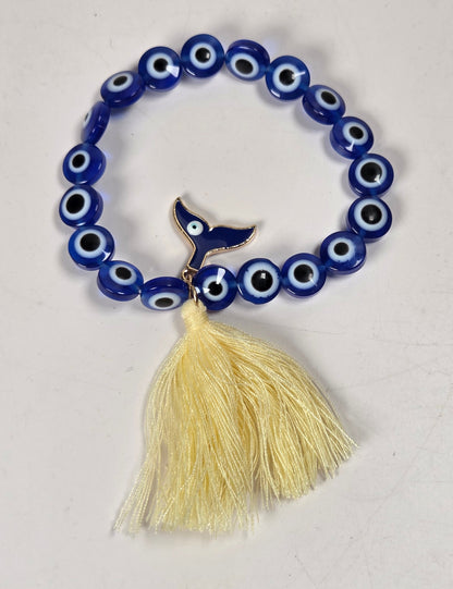 A blue beaded bracelet with evil eye charms and a dangling cream-colored tassel, photographed on a plain off-white surface.