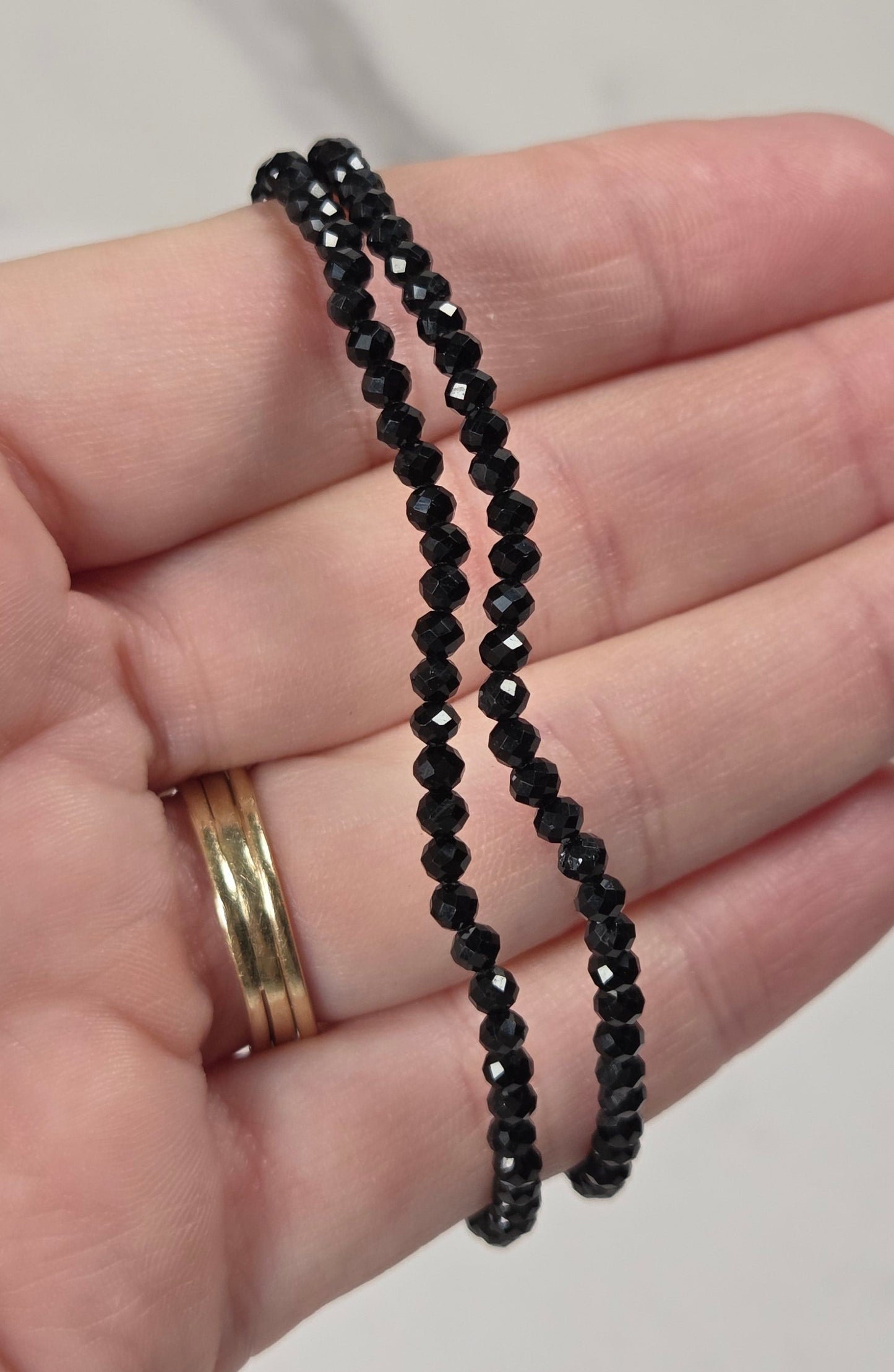 A close-up of a hand holding two thin strands of small black faceted beads, with a gold ring visible on one finger, against a light marble-patterned background.
