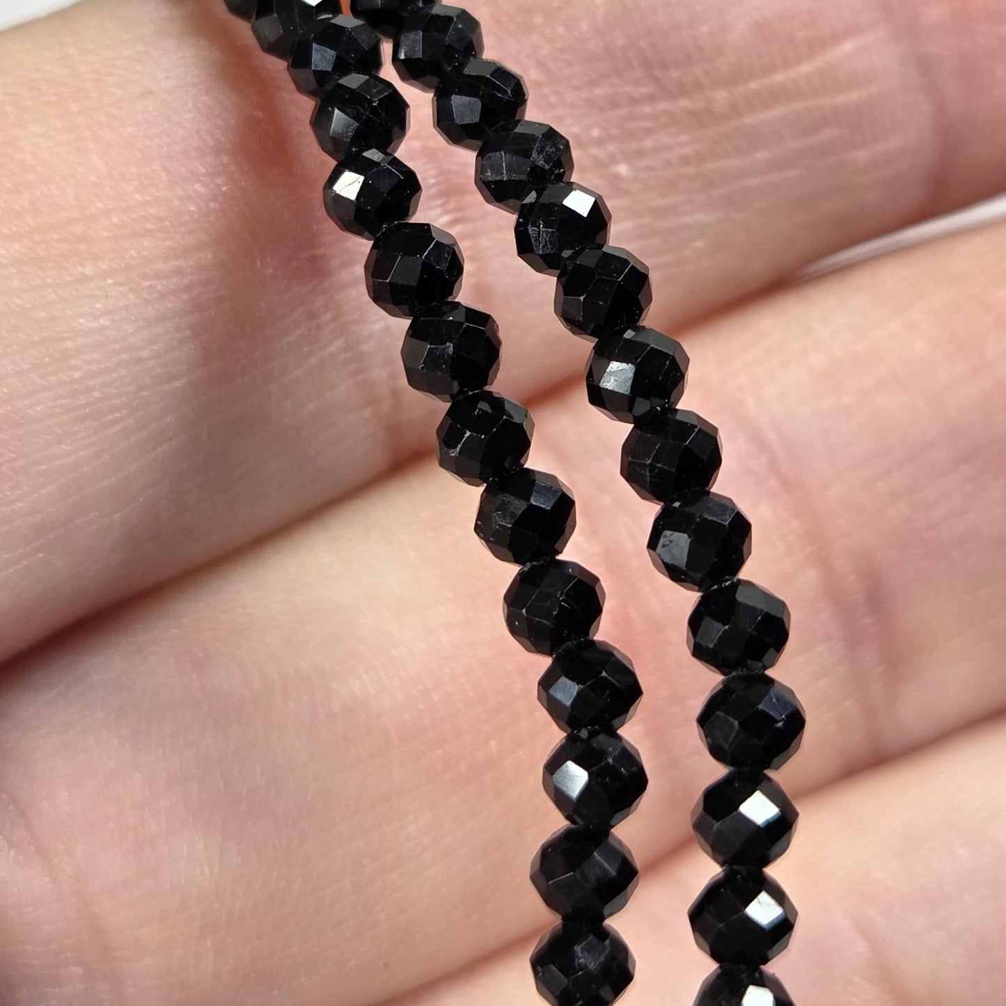A close-up of a hand holding a strand of faceted black beads, likely seed beads for jewelry making, against a soft-focus light gray background.