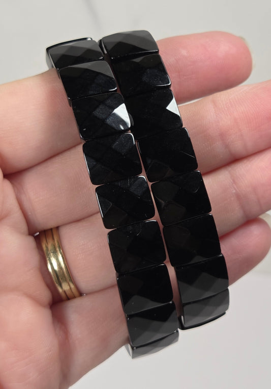 A pair of black faceted bead bracelets held in a person's hand, with a gold ring visible on the wrist. The background is a light marble-patterned surface.