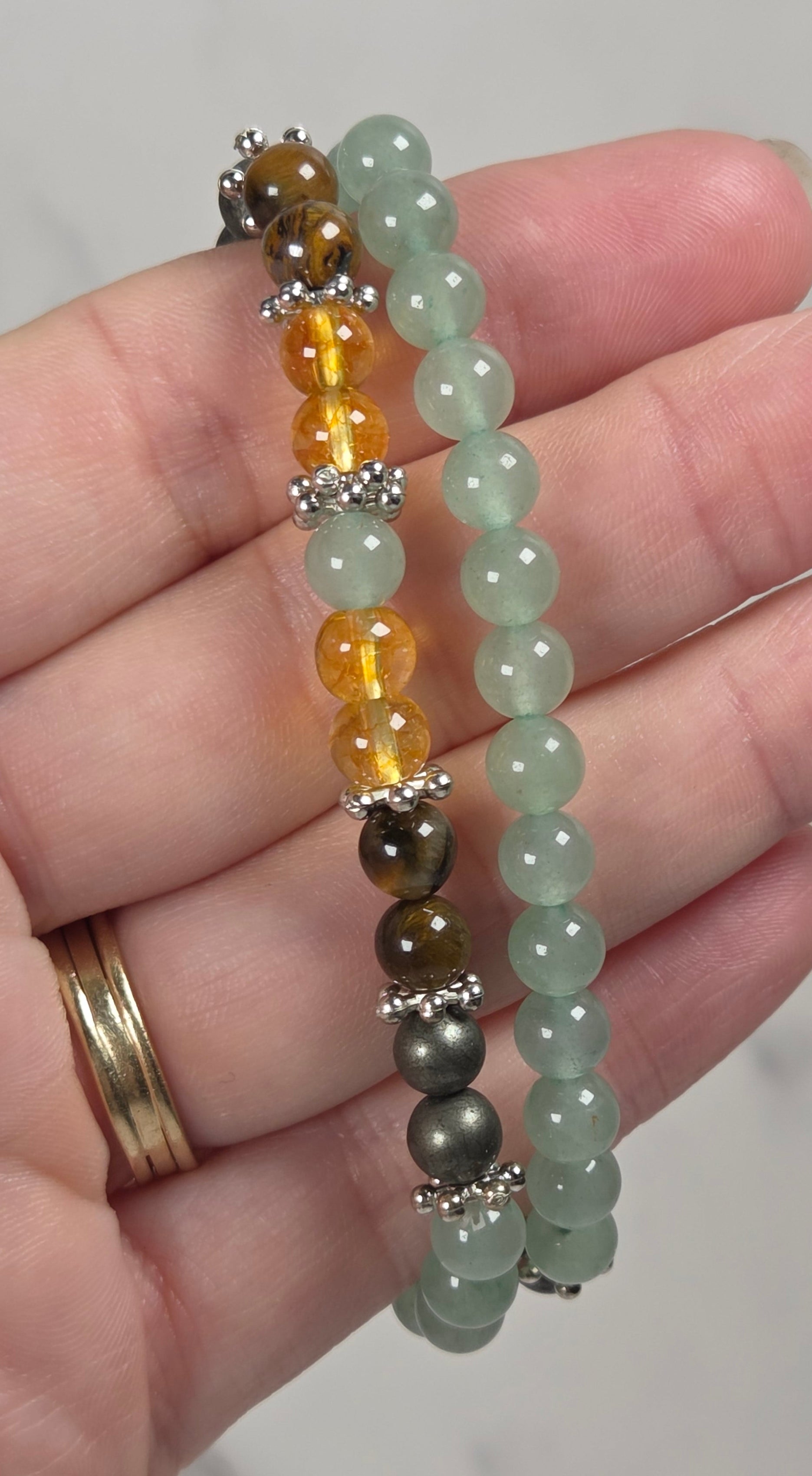 Two beaded stretch bracelets held in a hand. One bracelet features light green translucent beads with silver spacer accents, and the other features amber yellow, dark brown tiger's eye, and light green beads with silver accents.