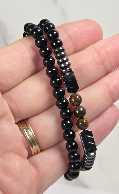A close-up of a hand holding two beaded bracelets. One bracelet is made of glossy black round beads, and the other features black faceted beads interspersed with brown tiger's eye round beads.