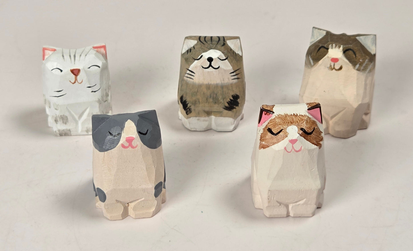 Five small cat figurines with a minimalist, geometric, origami-like style in various colors, arranged on a light beige background.