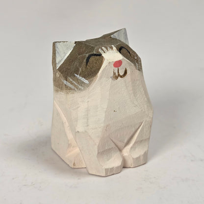 A small hand-carved wooden cat figurine with a painted gray and white head, black eyes and whiskers, and a small red nose, standing on a light gray surface.