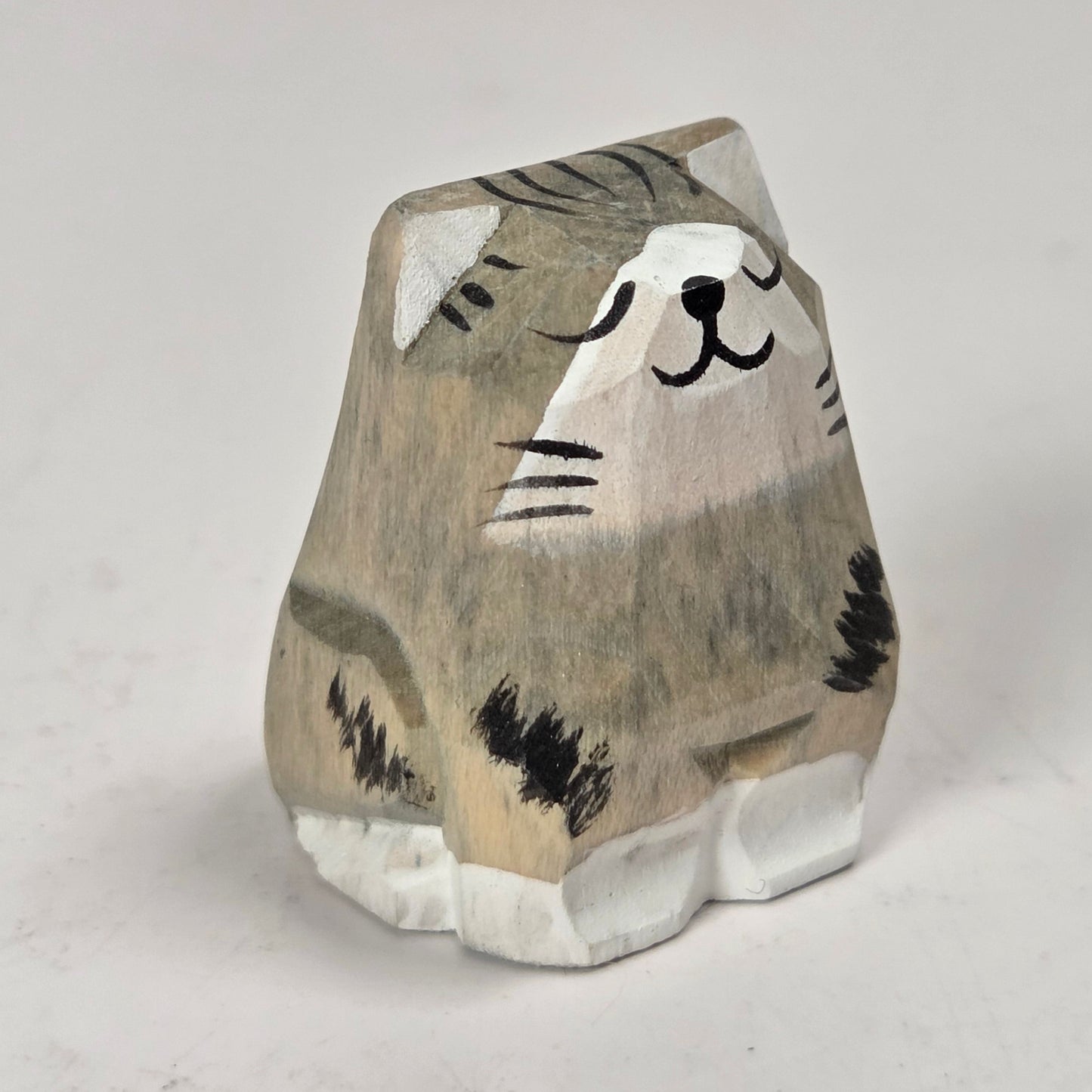 A small hand-carved wooden cat figurine with a gray and beige striped pattern, black painted details, and white paws, standing on a light gray background.