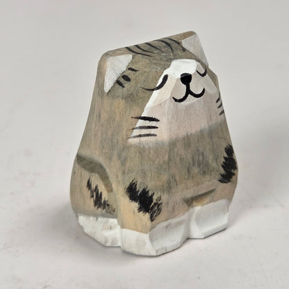 A small hand-carved wooden cat figurine with a gray and beige striped pattern, black painted details, and white paws, standing on a light gray background.