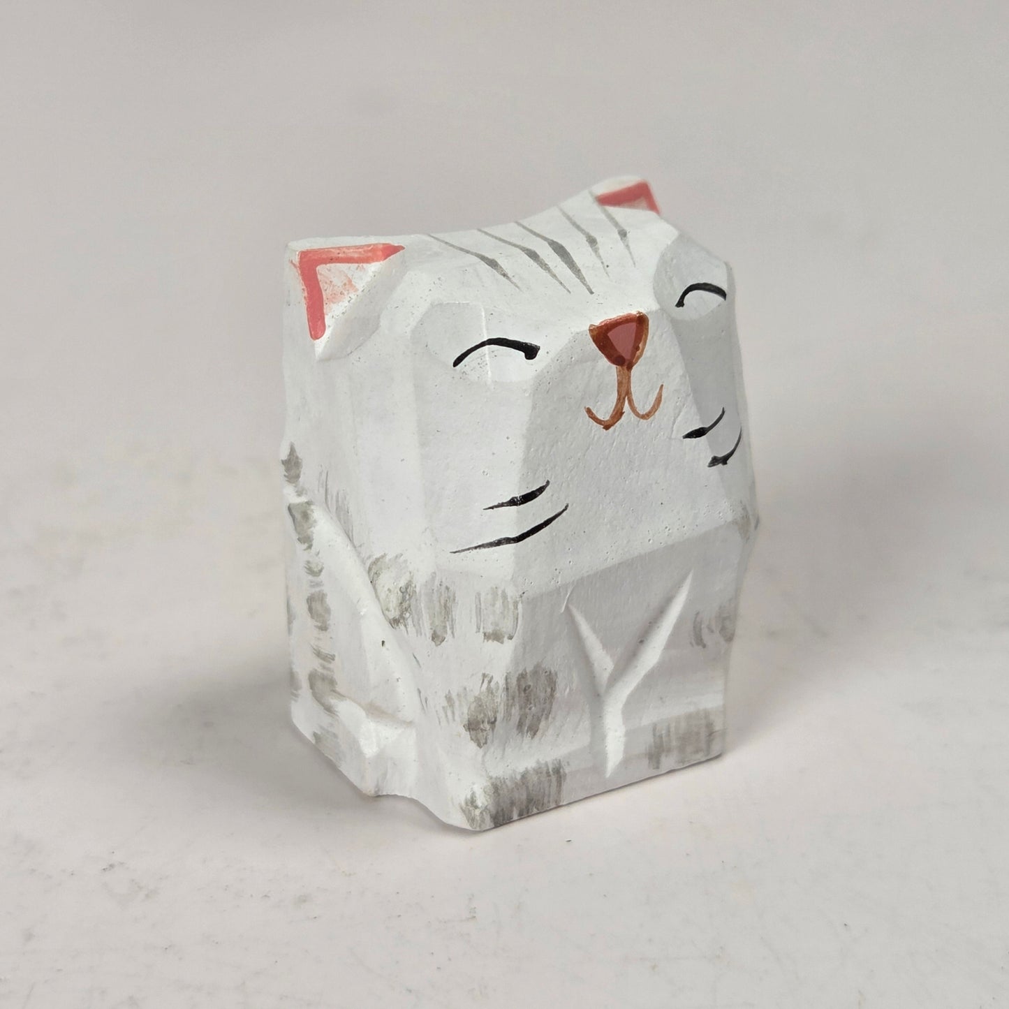 A small hand-carved cat figurine with a white-painted finish, featuring black painted facial details, gray carved wood grain, and pink inner ears, set against a neutral light gray background.
