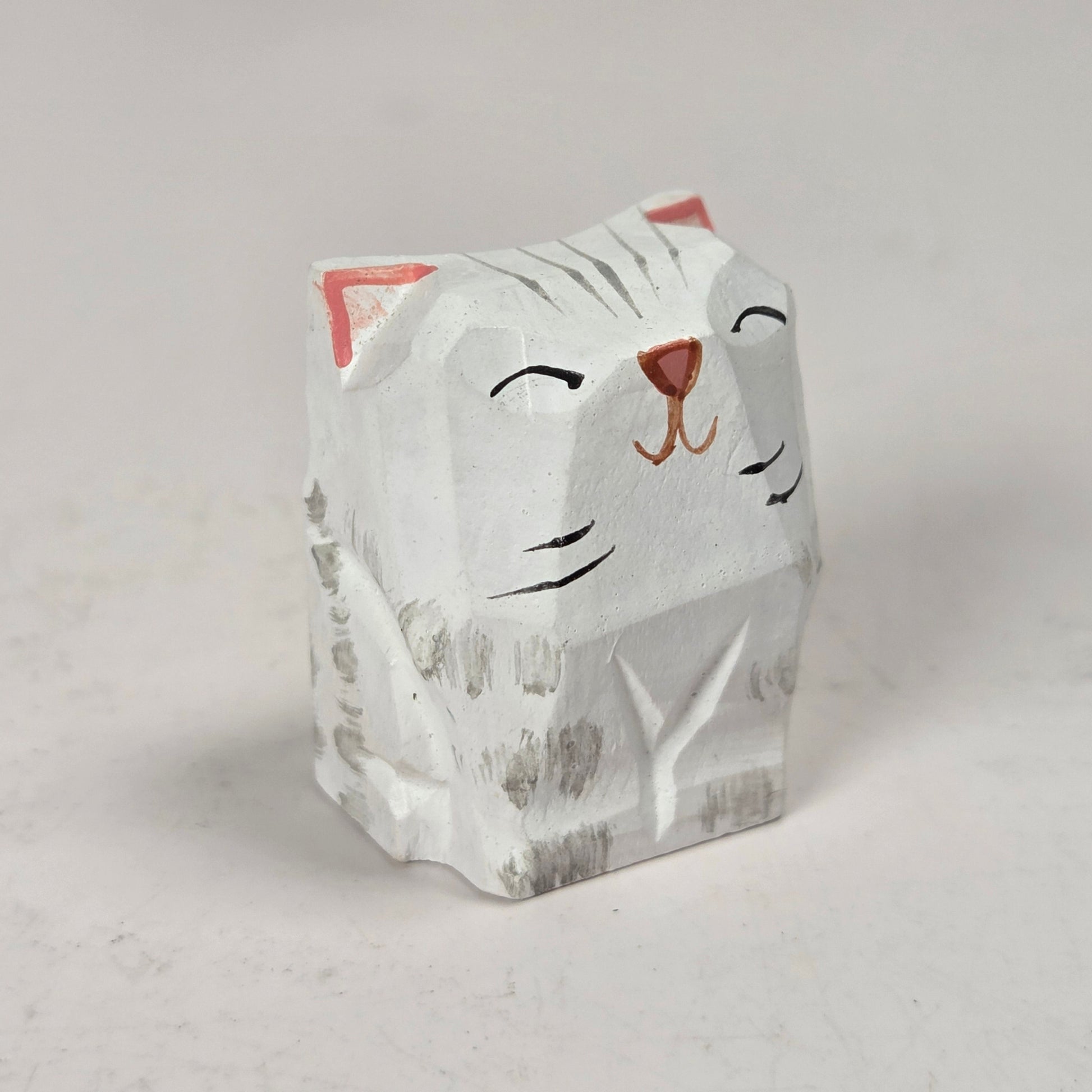 A small hand-carved cat figurine with a white-painted finish, featuring black painted facial details, gray carved wood grain, and pink inner ears, set against a neutral light gray background.