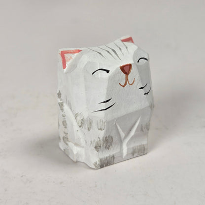 A small hand-carved cat figurine with a white-painted finish, featuring black painted facial details, gray carved wood grain, and pink inner ears, set against a neutral light gray background.
