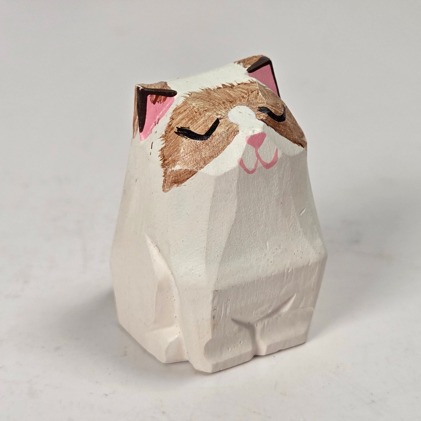 A small hand-carved wooden cat figurine with a white body, brown face, and pink inner ears and nose, shown on a light gray background.