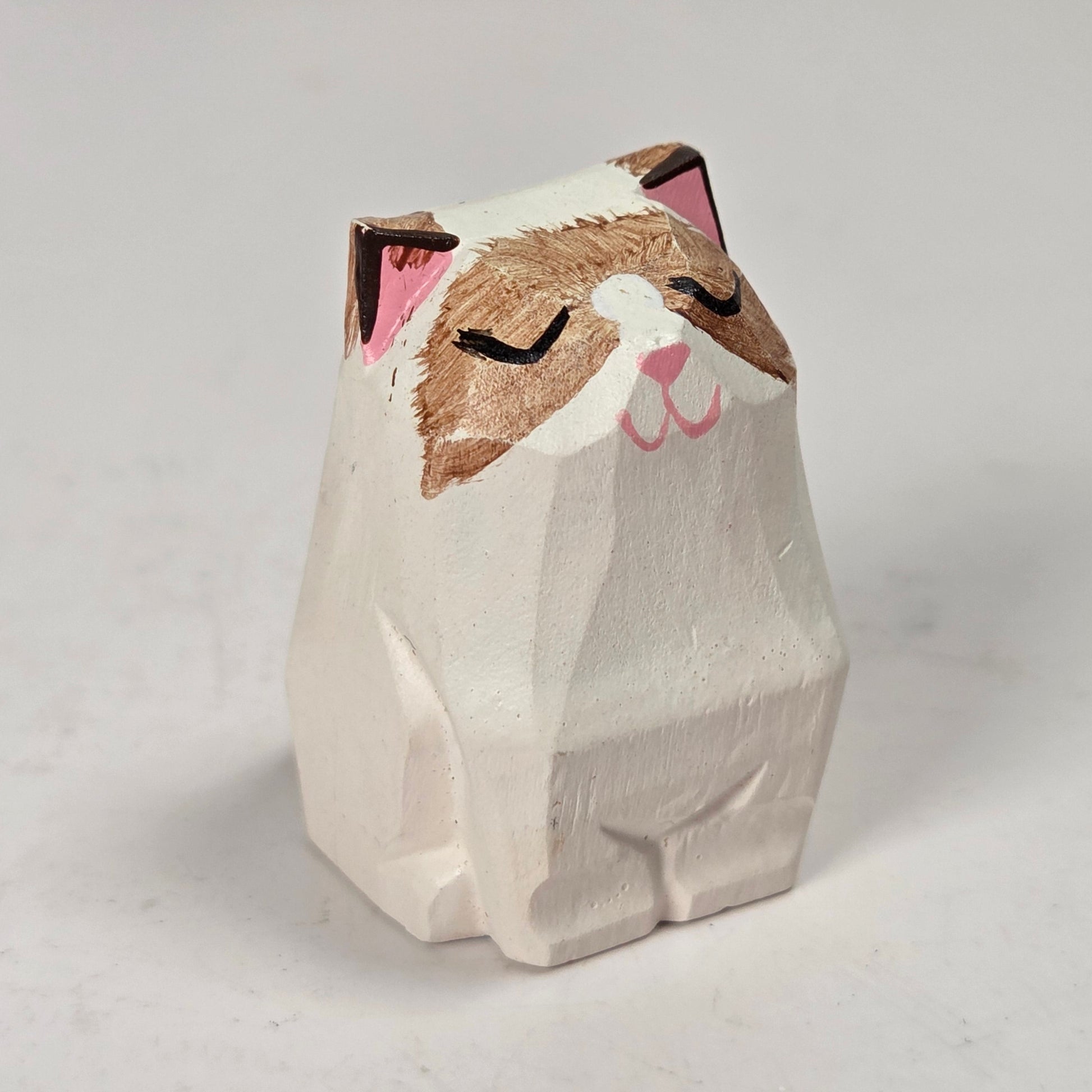 A small hand-carved wooden cat figurine with a white body, brown face, and pink inner ears and nose, shown on a light gray background.