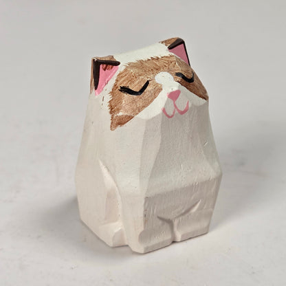 A small hand-carved wooden cat figurine with a white body, brown face, and pink inner ears and nose, shown on a light gray background.