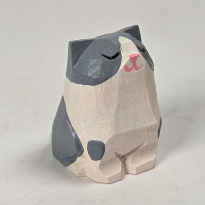 A small hand-carved wooden cat figurine with a gray and cream painted finish, featuring a pink nose and closed eyes, set against a light gray background.