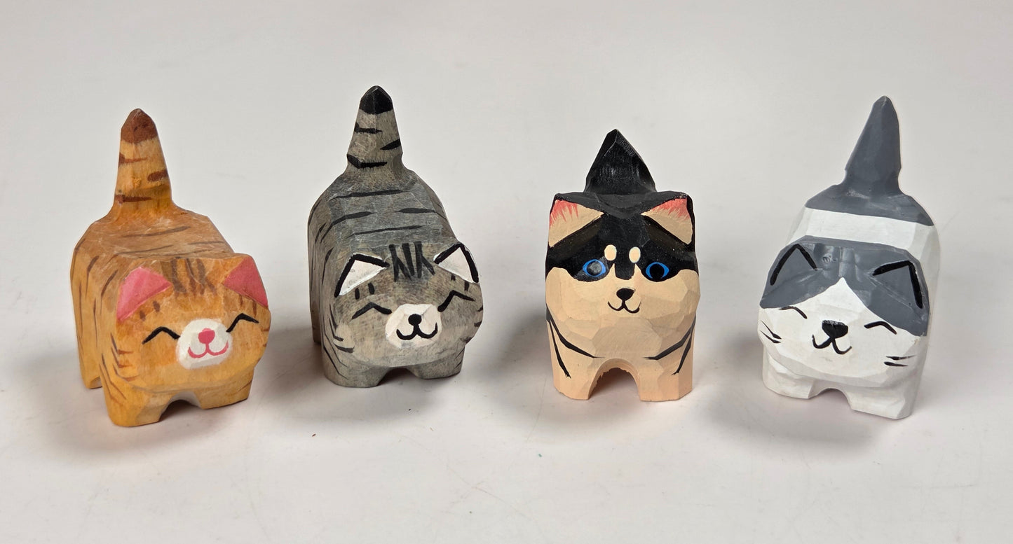 Four hand-carved wooden cat figurines in different colors and patterns, arranged in a row on a white surface with a marble-patterned background.
