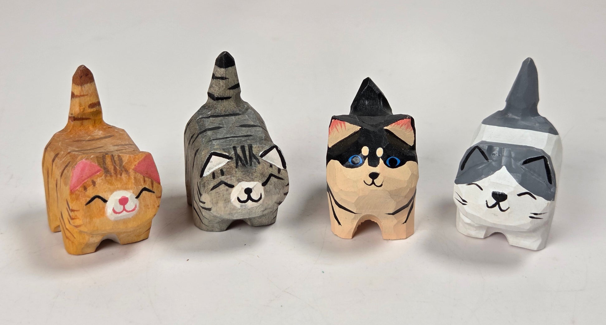 Four hand-carved wooden cat figurines in different colors and patterns, arranged in a row on a white surface with a marble-patterned background.