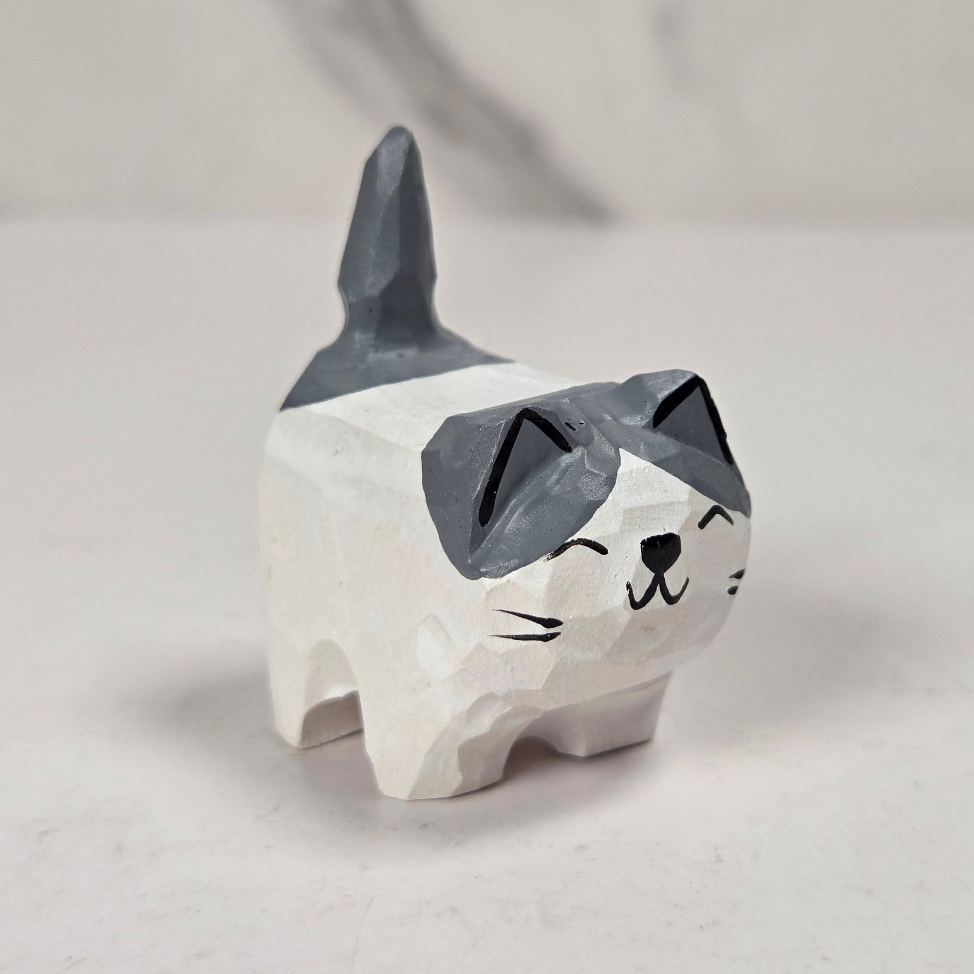 A small carved cat figurine with a white body and dark gray ears, eyes, and tail, standing on a light surface against a soft gray background.