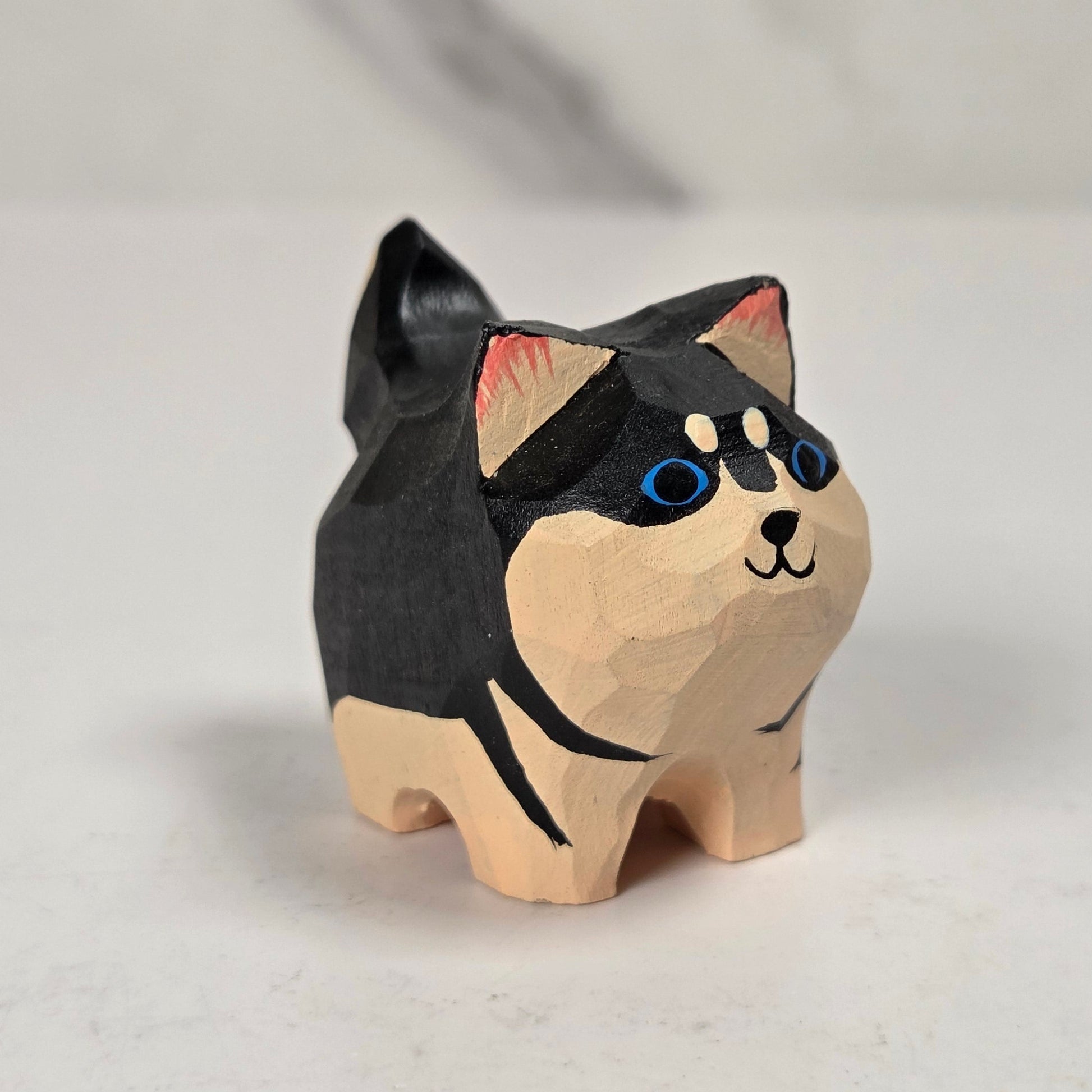 A small hand-carved wooden cat figurine with a black and beige body, blue painted eyes, and pink inner ears, standing on a light surface.