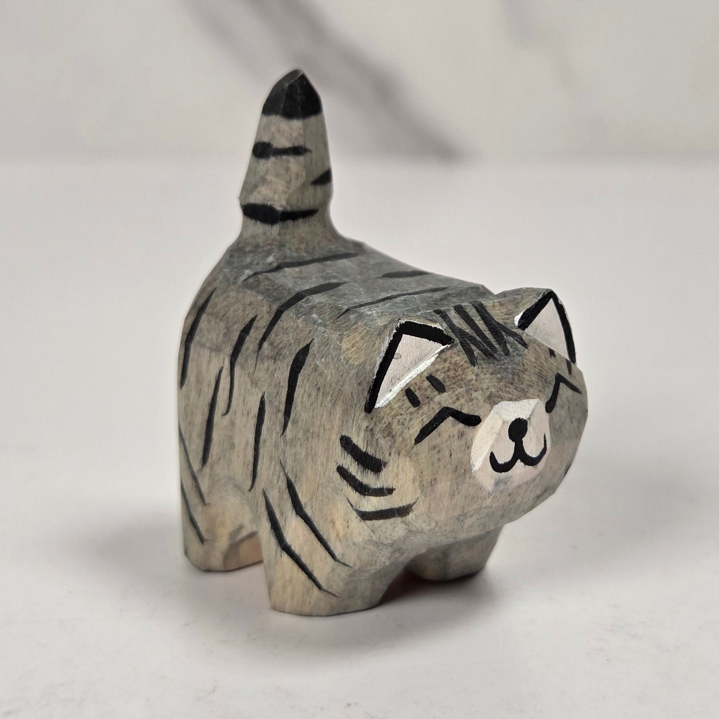 A small carved cat figurine with a gray and black striped pattern, standing on a light gray surface against a white marble-look background.