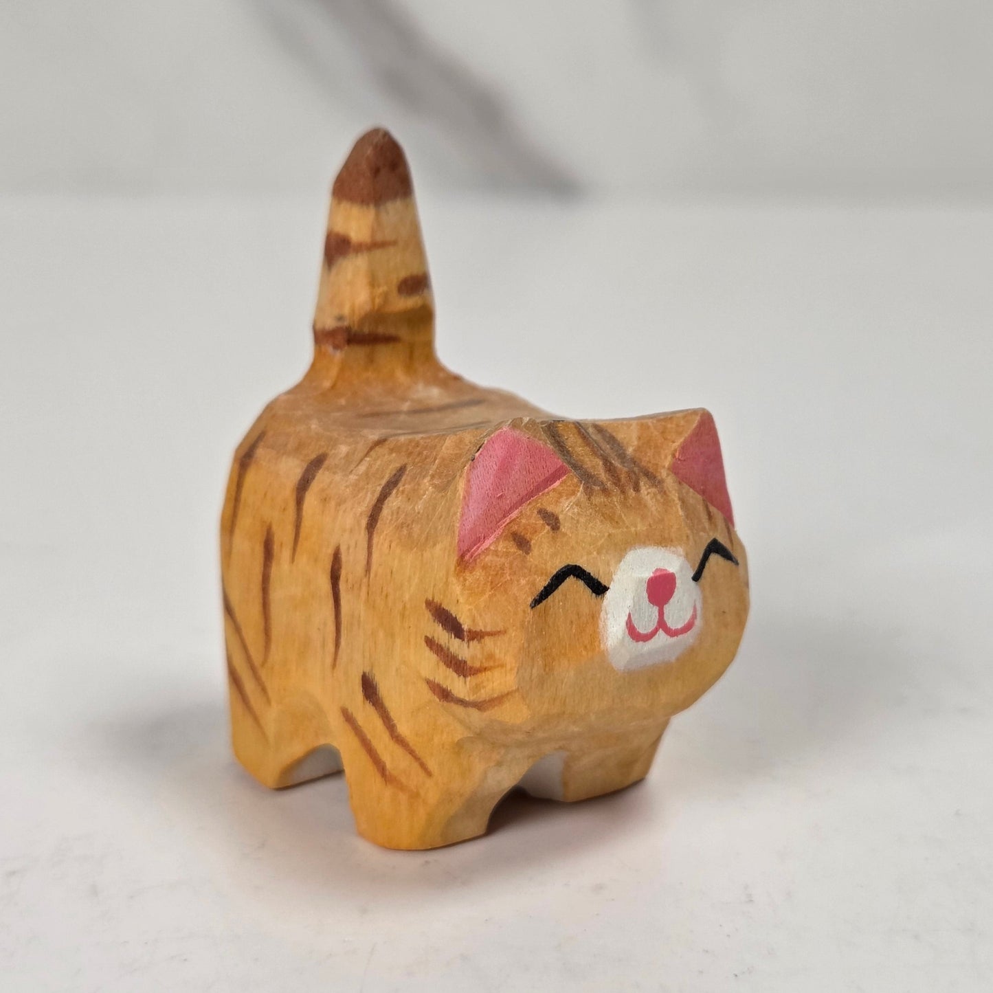 A small hand-carved wooden cat figurine with orange and brown stripes, pink inner ears, and a white face, standing on a light surface.