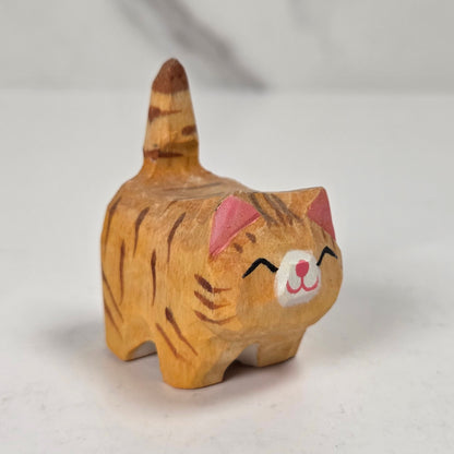 A small hand-carved wooden cat figurine with orange and brown stripes, pink inner ears, and a white face, standing on a light surface.