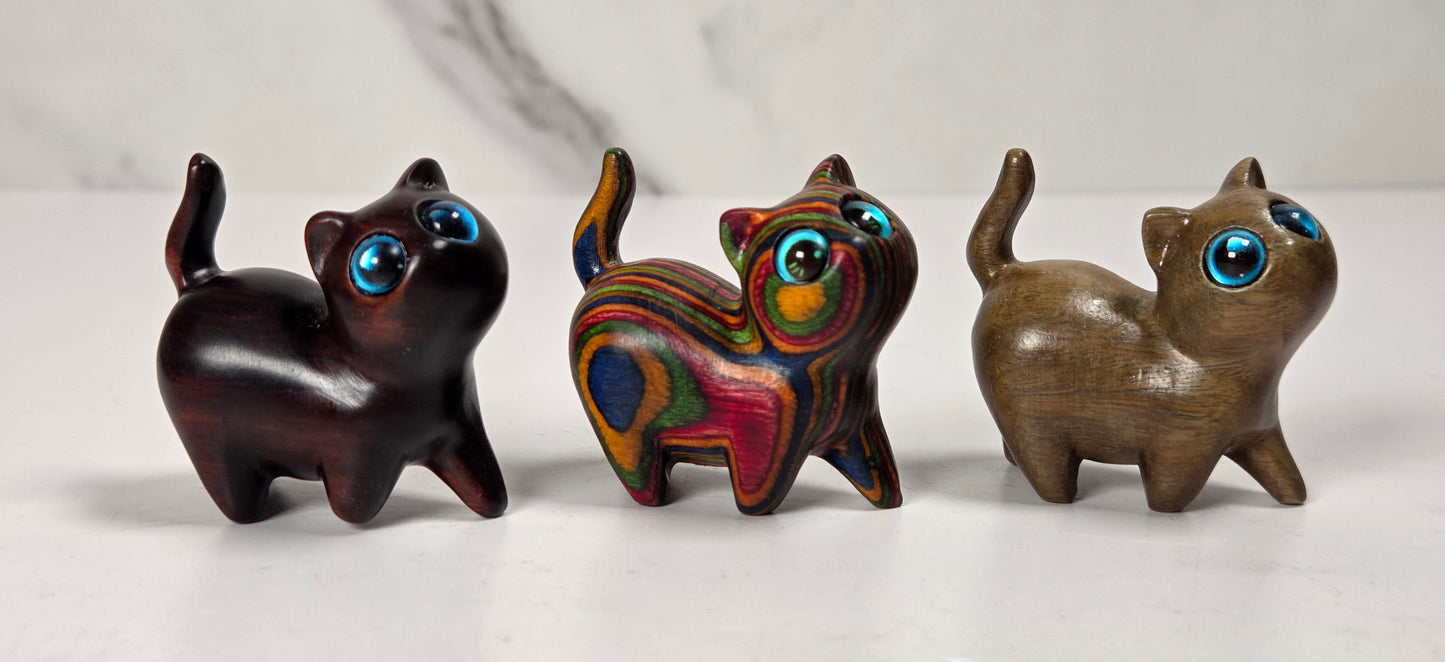 Three small carved cat figurines with large blue eyes, displayed side by side on a light marble surface against a white marble background.
