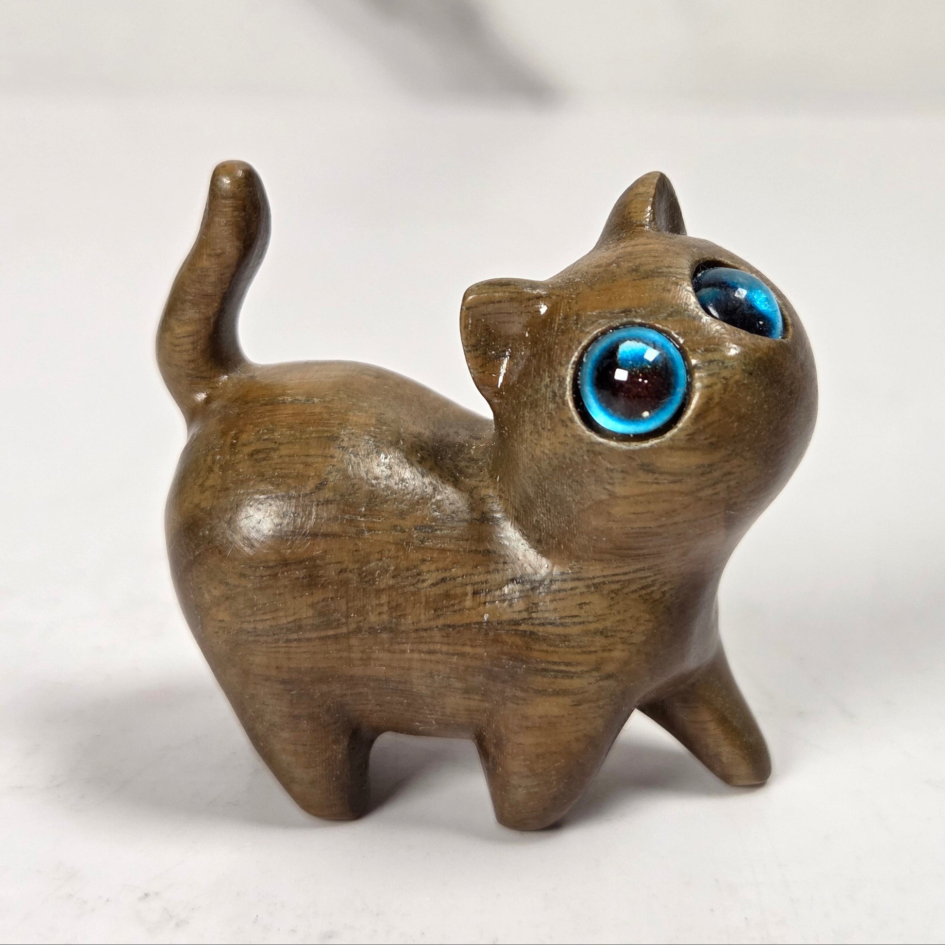 A small wooden cat figurine with a glossy brown finish and large, expressive blue glass eyes, standing on a light marble-like surface.