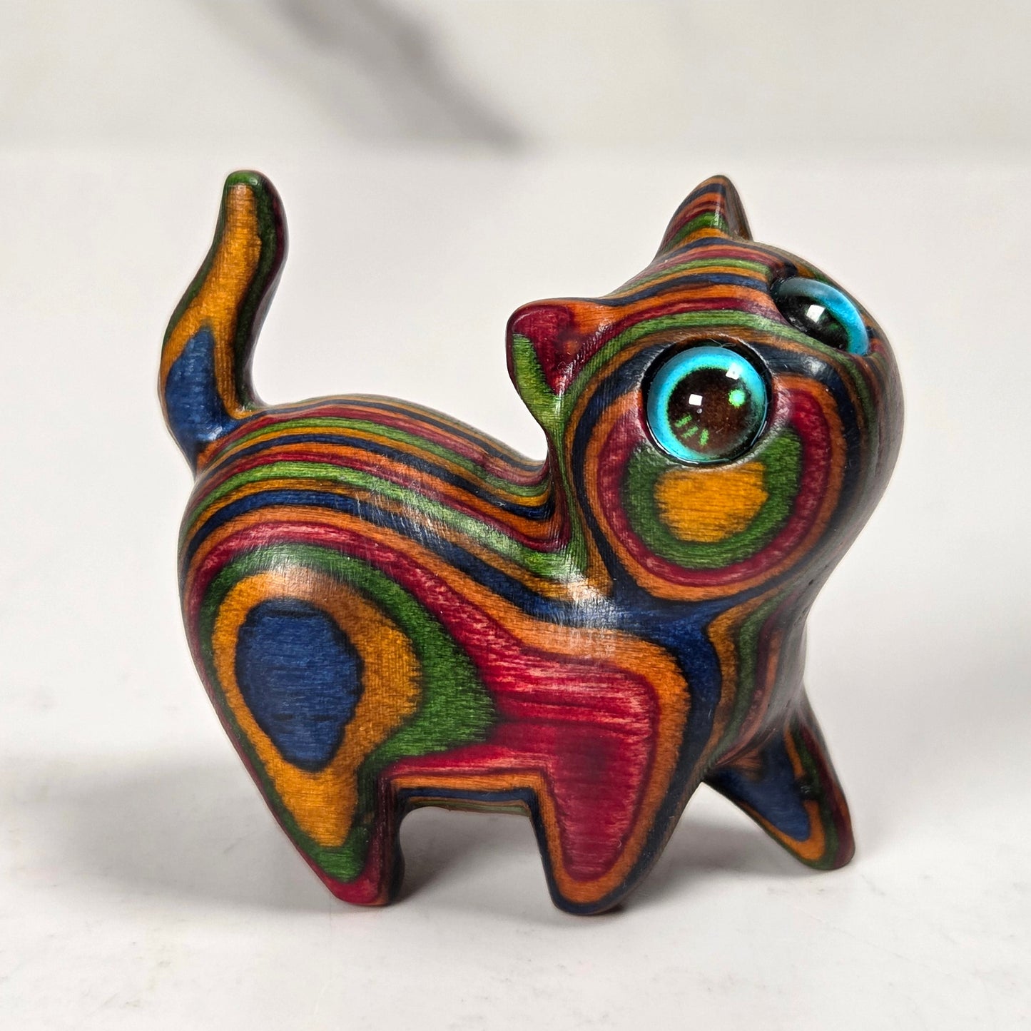 A small handcrafted wooden cat figurine with vibrant, swirling multicolored stripes of red, green, yellow, blue, and orange, featuring large glossy blue eyes, standing on a light gray marble surface.