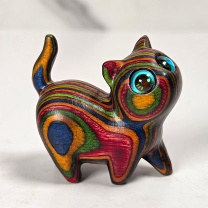 A small handcrafted wooden cat figurine with vibrant, swirling multicolored stripes of red, green, yellow, blue, and orange, featuring large glossy blue eyes, standing on a light gray marble surface.